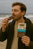 Man on a beach eating dark chocolate with a bar labelled 'Tasman Sea Salt' in his other hand.