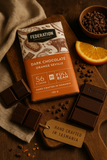 A bar of Federation Dark Chocolate Orange Seville placed on a wooden surface, surrounded by broken pieces of chocolate, an orange slice, and chocolate chips. A tag with 'HAND CRAFTED IN TASMANIA' is also visible.