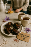 Chocolate treats on a plate with a tag labelled 'Tasmanian Saffron Frogs' on a table with cups of tea and flowers.